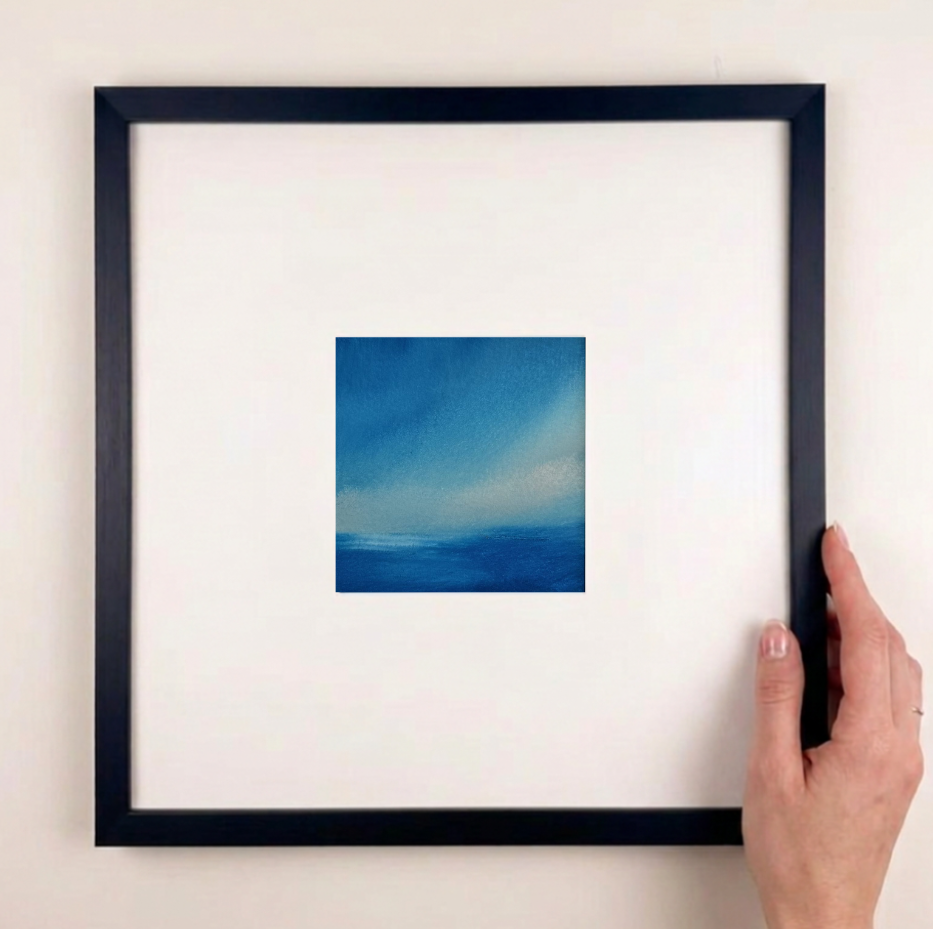 A hand holding a black square frame with a wide white mat, featuring a small blue abstract painting in the center, against a plain off-white wall.