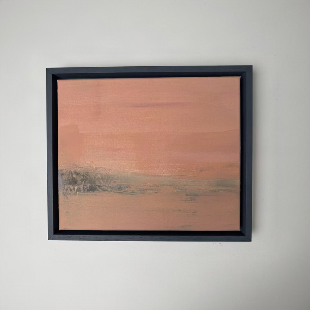 A framed abstract landscape painting featuring soft horizontal brushstrokes in dusty pink, terracotta, and muted gray-green tones, housed in a dark charcoal frame against a plain white wall.