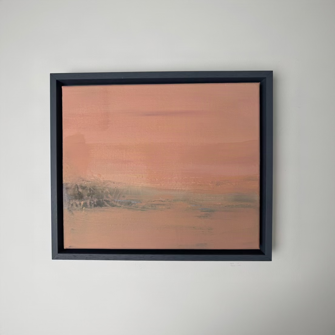 A framed abstract landscape painting featuring soft horizontal brushstrokes in dusty pink, terracotta, and muted gray-green tones, housed in a dark charcoal frame against a plain white wall.