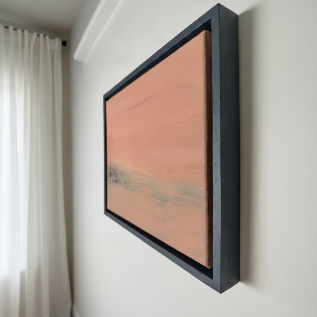 A framed abstract landscape painting featuring a warm peach and pink sky with a muted gray horizon, housed in a thick black frame and shown hanging on a white wall.
