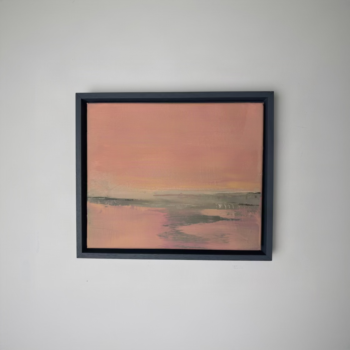 A framed abstract landscape painting featuring a soft pink and peach sky over muted green and gray land, housed in a dark charcoal frame and mounted on a plain white wall.