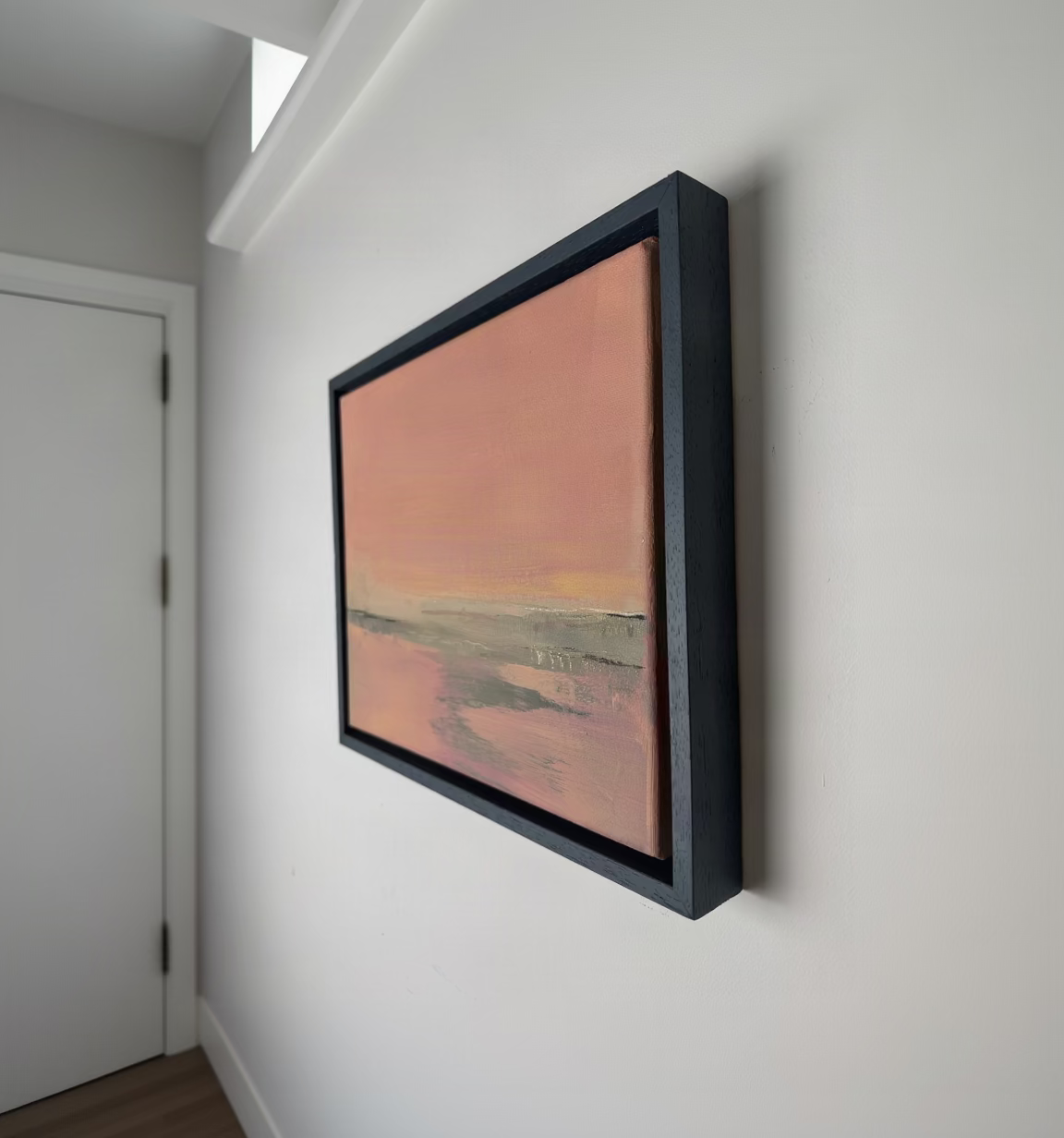 A framed abstract landscape painting hangs on a white wall in a hallway. The artwork features a soft pink and peach sky with muted gray-green and beige tones below, enclosed in a dark charcoal frame.